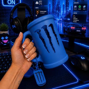 May include: A matte blue beer mug featuring the iconic three-claw scratch logo cut out on the side. The mug includes a matching blue dog tag style charm attached to the handle by a small ball chain, creating a cohesive look for a gaming setup.