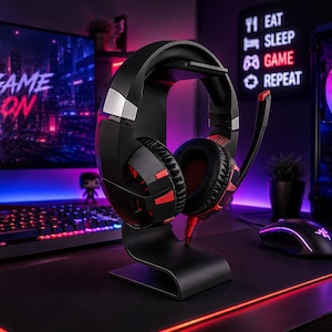 May include: Black and red gaming headset on a black stand. The headset has a microphone and is positioned in front of a computer monitor displaying "GAME ON". A neon sign reads "EAT SLEEP GAME REPEAT".