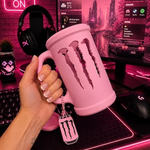 May include: A light pink cylindrical mug featuring a cutout of the three-clawed Monster Energy logo. A small matching pink tag with a black Monster Energy logo hangs from the handle by a beaded chain. The mug has a smooth, matte finish.