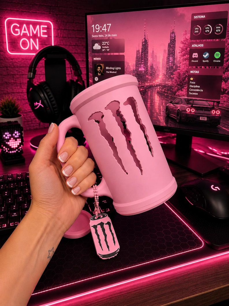 May include: A light pink cylindrical mug featuring a cutout of the three-clawed Monster Energy logo. A small matching pink tag with a black Monster Energy logo hangs from the handle by a beaded chain. The mug has a smooth, matte finish.