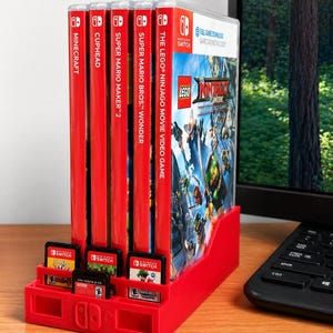May include: A red plastic Nintendo Switch game organizer holding several game cases, including "Minecraft", "Cuphead", and "Super Mario Maker 2". The organizer also has slots for game cartridges. The background includes a computer monitor and keyboard.