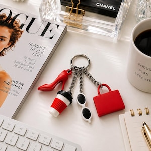May include: A silver keychain with charms shaped like a red high heel, a coffee cup, sunglasses, and a handbag. A Vogue magazine is in the background, with the text "SUMMER STYLE EDIT". A white mug with the words "FOCUS PLAN EXECUTE" is also visible.