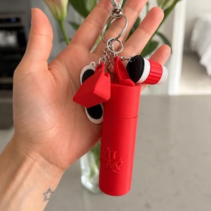 May include: A red keychain with multiple charms, including a small shopping bag, sunglasses, and a coffee cup. The keychain also features a cylindrical container with the text "VIL" and a silver clasp.