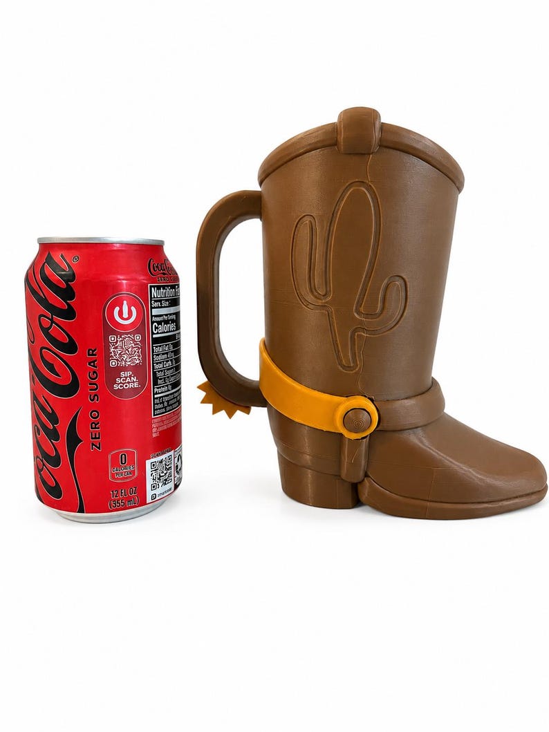 May include: A brown cowboy boot-shaped mug with a handle, featuring a raised cactus design on the side and a yellow spur attached to the heel. A red 12 fluid ounce Coca-Cola Zero Sugar can is placed next to the mug for a size comparison.