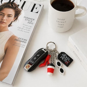 May include: A black car key with a Porsche logo, attached to a keychain with charms including a red high heel, a coffee cup, sunglasses, and a tag that says "The Devil Wears Prada." A white mug with "Good Things Take Time" is nearby.