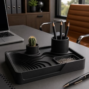 May include: A matte black desk organizer tray featuring a topographical, layered design. It includes a small cylindrical pot containing a green cactus, a larger cylindrical pencil holder with three black pencils, and a rectangular compartment for paper clips.