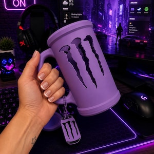 May include: A matte lavender mug featuring a large, vertical, three-clawed cutout design on the front. A small, matching lavender-colored metal tag shaped like a beverage can with the same three-clawed logo hangs from the handle by a chain.