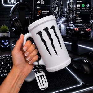 May include: A white ceramic mug featuring a large, vertical, claw-mark cutout design on the front. A small, black, rectangular metal tag with a white claw-mark logo hangs from the handle by a black beaded chain, matching the mug's aesthetic.