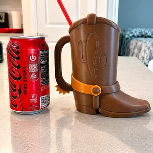May include: A brown cowboy boot-shaped mug with a handle, a yellow spur, and a red straw, positioned next to a 12 fluid ounce Coca-Cola Zero Sugar soda can for scale. The mug features an embossed cactus design on the side and a yellow strap detail.