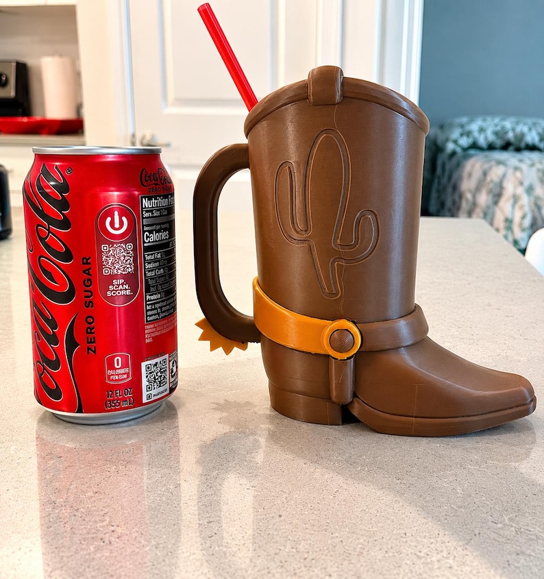 May include: A brown cowboy boot-shaped mug with a handle, a yellow spur, and a red straw, positioned next to a 12 fluid ounce Coca-Cola Zero Sugar soda can for scale. The mug features an embossed cactus design on the side and a yellow strap detail.