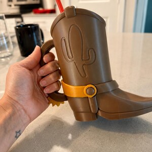 May include: A brown plastic cup shaped like a cowboy boot with a handle, featuring a raised cactus design on the side, a yellow spur at the heel, a yellow strap detail across the ankle, and a red straw protruding from the top opening.