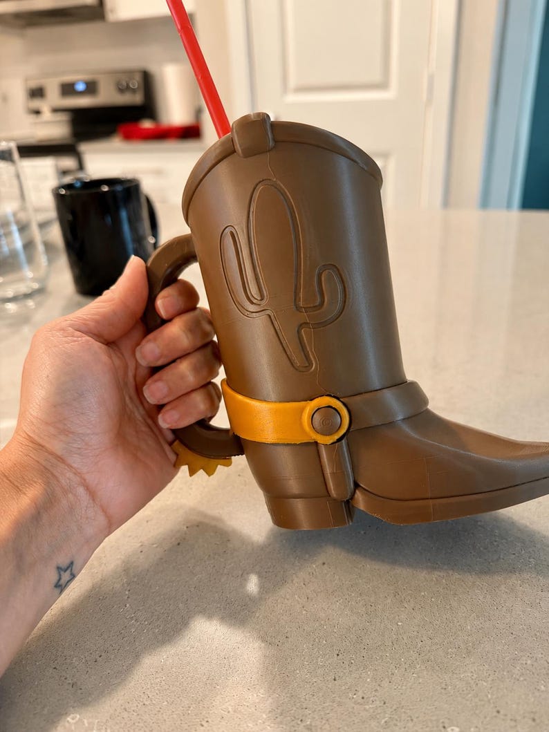 May include: A brown plastic cup shaped like a cowboy boot with a handle, featuring a raised cactus design on the side, a yellow spur at the heel, a yellow strap detail across the ankle, and a red straw protruding from the top opening.