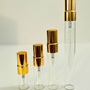 May include: Four clear glass perfume atomizers with gold-colored caps are arranged in a row. The atomizers are labeled with their sizes: 2ml, 3ml, 5ml, and 10ml. The background is a plain white.