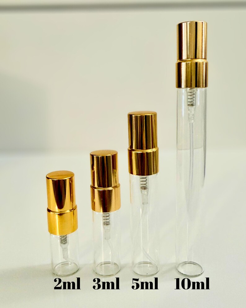 May include: Four clear glass perfume atomizers with gold-colored caps are arranged in a row. The atomizers are labeled with their sizes: 2ml, 3ml, 5ml, and 10ml. The background is a plain white.