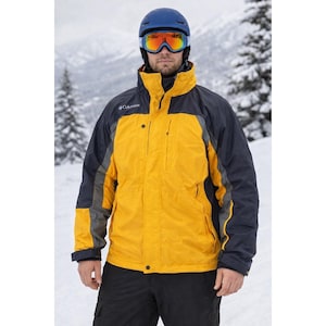 May include: A yellow and navy blue winter jacket with a high collar and multiple pockets. The jacket has grey accents and the Columbia logo on the chest. The person is wearing a blue helmet and orange-tinted ski goggles. The background is a snowy mountain scene.