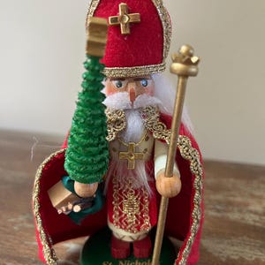 Op de afbeelding: Een houten notenkrakerfiguur van Sint Nicolaas met een rode mijter met een gouden kruis, een rode mantel met gouden rand en een wit gewaad. Het figuur houdt een groene boom en een gouden staf vast op een groene voet met de tekst St. Nicholas 1650.