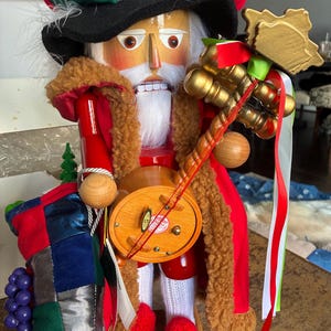 May include: A wooden nutcracker doll in a festive outfit. The doll has a white beard, a red coat with a brown fur collar, and a tall hat with red, white, and green accents. It holds a musical instrument and a decorative staff.