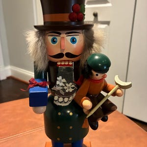 Könnte beinhalten: Ein Nussknacker-Figur aus Holz mit Zylinder, Fellbesatz und grünem Mantel. Der Nussknacker hält ein kleines Geschenk und eine kleinere Figur. Die Figur hat ein detailliertes Gesicht und ist in verschiedenen Farben bemalt.