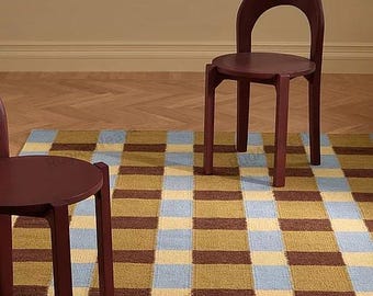 May include: A modern interior scene featuring a checkered rug in shades of brown, blue, and yellow. Two burgundy chairs with rounded backs and circular seats are positioned on the rug. A woven wall hanging with brown fringe hangs in the background.