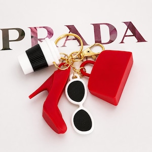 May include: A gold-toned keychain featuring miniature charms: a red high-heeled shoe, a red handbag, a white and black coffee cup, white sunglasses with black lenses, and a gold-toned ring. The word "PRADA" is visible in the background.