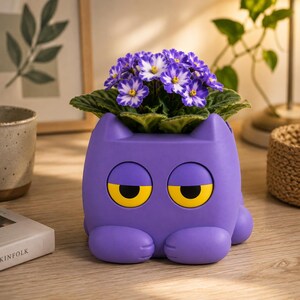 Op de afbeelding: Een paarse katvormige plantenpot met twee grote gele ogen en afgeronde oortjes. De pot bevat een Afrikaans viooltje met donkergroene bladeren en kleine paars-witte bloemen. Het katfiguur heeft twee voorpoten die naar voren rusten.