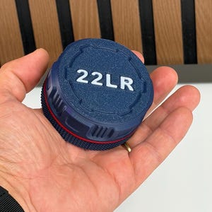 May include: A circular, dark blue container lid featuring the white text 22LR embossed on the top surface. The lid has a textured, knurled side grip with a thin red accent line running horizontally around the circumference of the cylindrical edge.