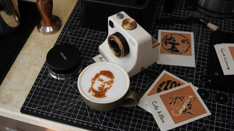 May include: A white digital camera with a gold lens ring and top dial sits next to a ceramic mug filled with coffee featuring a portrait of a person in the foam. Nearby are several square photo prints with graphic designs and the text Cute kitten.