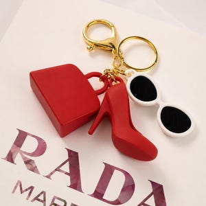 Puede incluir: Un llavero dorado con accesorios rojos: un mini bolso, un zapato de tacón y gafas de sol blancas con lentes negras. El llavero está sobre una superficie blanca con el texto "RADA MARFA".