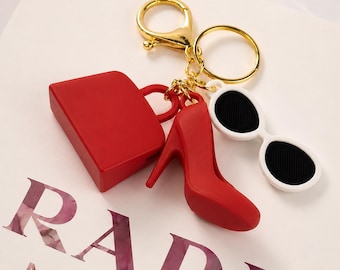 Puede incluir: Un llavero dorado con accesorios rojos: un mini bolso, un zapato de tacón y gafas de sol blancas con lentes negras. El llavero está sobre una superficie blanca con el texto "RADA MARFA".