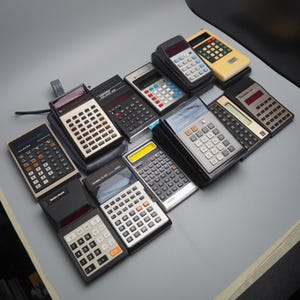 May include: A selection of retro calculators in shades of black, silver, and cream. The calculators feature diverse button arrangements and display screens. Some are in protective cases, and all seem to be from the 1970s or 1980s.