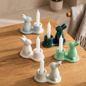 Op de afbeelding: Een verzameling keramische konijnenkaarsenhouders in verschillende pastelkleuren, elk met een witte kaars. De konijnenfiguren zijn gecombineerd met een kaarsenhouder op een kleine, ovale basis. Geplaatst op een houten oppervlak.