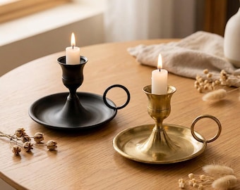 May include: Two metal candle holders with circular bases and side ring handles. One holder is matte black and the other is brushed brass. Each features a central cup holding a white taper candle with a lit flame, creating a balanced display.