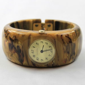 May include: A gold-toned wristwatch with a brown and yellow marbled plastic bangle band. The watch face is round and has black hands and black Roman numeral markings. The watch face says "Geneva" at the top.