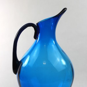 May include: A cobalt blue glass pitcher with a curved handle. The pitcher has a rounded body and a wide mouth.