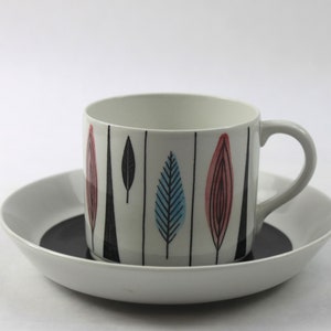 May include: White ceramic mug and saucer set. The mug features a gray handle and is decorated with vertical black lines and colorful leaf-like designs in red, blue, and black. The saucer has a white rim and a dark gray center.