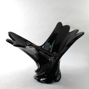 May include: A glossy black decorative bowl with a unique, organic shape. The bowl has a flared, petal-like rim and a twisted, sculptural base. The surface reflects light, creating highlights and shadows. The bowl's design is modern and artistic.