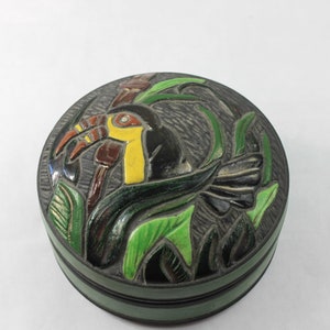May include: A round ceramic trinket box with a toucan design. The toucan is painted in yellow, red, and black, and is surrounded by green leaves. The box is black with a green rim.