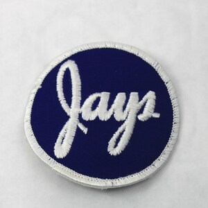 May include: A circular embroidered patch featuring a deep navy blue background with the word Jays stitched in white cursive script. The patch has a white merrowed border edge that frames the circular design, creating a clean and finished look.