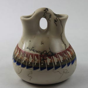 May include: A cream-colored ceramic vase with a unique shape, featuring a central opening and two side handles. The vase is adorned with a colorful geometric pattern in red, blue, and brown. Black lightning-like lines decorate the surface.