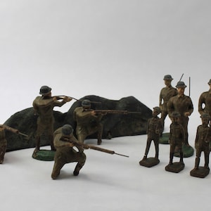 May include: A collection of vintage toy soldiers in various poses, including kneeling and standing, with rifles. The figures are painted in shades of brown and green, with black hats. A dark green, textured terrain piece is in the background.