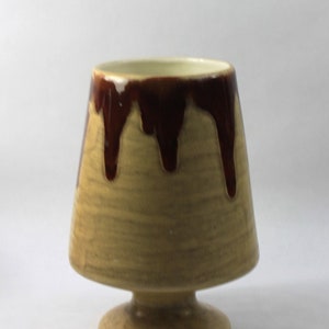 May include: A ceramic planter with a brown glaze and a light brown base. The planter has a wide, flared top and a narrow base.