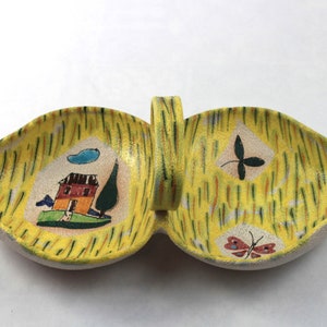 May include: A ceramic serving dish with two leaf-shaped compartments and a handle. The dish features a yellow background with vertical brushstrokes and painted illustrations of a house, tree, cloud, and butterflies. The dish has a light gray rim.