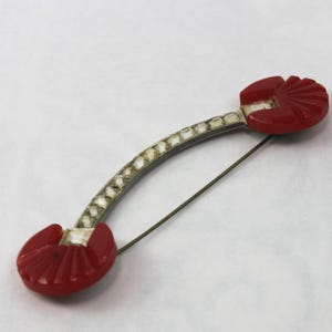 May include: A vintage brooch with a curved design. The brooch features two red, fan-shaped accents at each end, connected by a silver-toned, curved bar with small, clear embellishments. A pin extends from the back for securing the brooch.