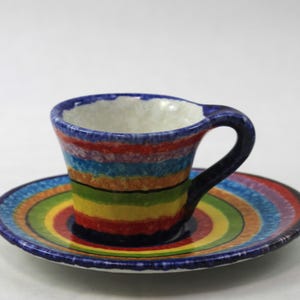 May include: A colorful ceramic teacup and saucer set. The cup and saucer are decorated with horizontal bands of rainbow colors, including red, orange, yellow, green, blue, and purple. The cup has a dark blue handle and rim.