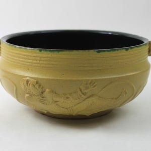 May include: A handcrafted ceramic bowl with a light yellow exterior and a black interior. The bowl features detailed relief carvings and two decorative handles. The rim has a green accent.