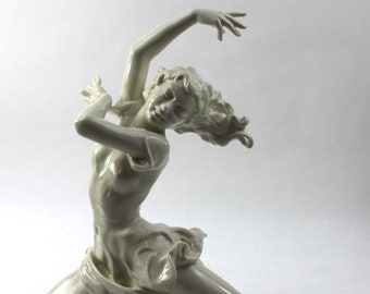 May include: A white porcelain figurine of a woman in a flowing dress, posed in a dynamic dance move with her arm raised and her head tilted back.