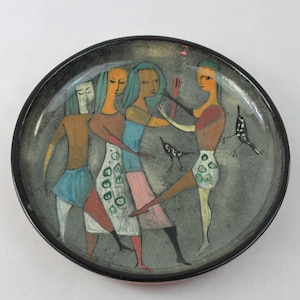 May include: A decorative ceramic plate with a black rim and a gray interior. The plate features a stylized painting of figures in dresses and birds. The figures are in various colors, including orange, blue, and pink.