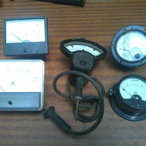 May include: A selection of antique electrical meters, including ammeters and voltmeters, with black and white dials and needles. The meters are in different shapes and sizes, with a rectangular ammeter and a circular voltmeter.