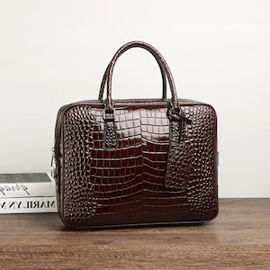 May include: A brown leather briefcase with a crocodile skin texture. The bag has a top handle, a rectangular shape, and a matching luggage tag. The briefcase is sitting on a wooden surface.