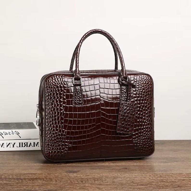 May include: A brown leather briefcase with a crocodile skin texture. The bag has a top handle, a rectangular shape, and a matching luggage tag. The briefcase is sitting on a wooden surface.